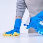 Young woman housekeeper is doing cleaning white table in apron with blue gloves, spray cleaner, wet yellow rag, close up, copy space, blank design concept.